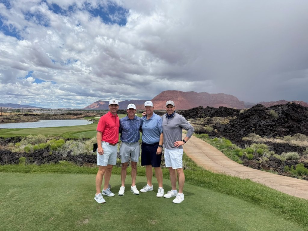 The group of four golfers on a tee box with black lava rock and red cliffs behind them at Black Desert Resort, Ivins, Utah