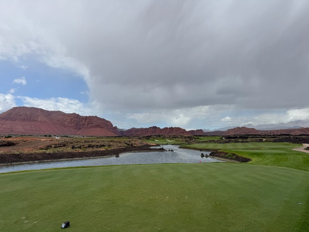 View from a tee box over a pond and fairway with black lava rock and red cliffs at Black Desert Resort, Ivins, Utah